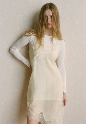 Junge Frau mit langen blonden Haaren, die ein cremefarbenes, mit Spitze besetztes Unterkleid über einem weißen Langarmshirt trägt und vor einer neutralen Wand steht.