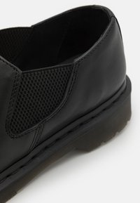 Scarpa in pelle nera con un pannello in rete testurizzato, forma arrotondata, suola leggermente elevata e cuciture visibili.