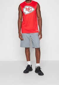 Maillot rouge sans manches avec un logo des Kansas City Chiefs, short de sport gris et baskets noires avec des accents blancs.