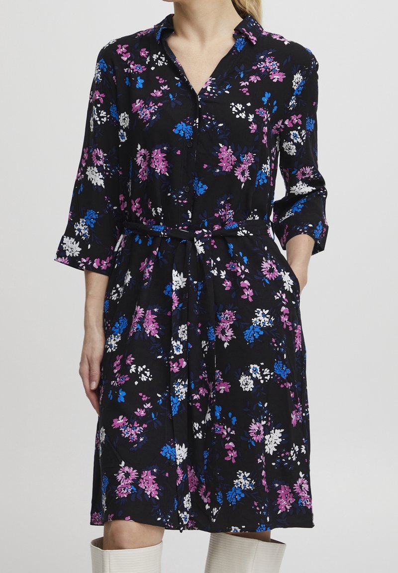 Femme portant une robe noire à fleurs roses, bleues et blanches, manches trois-quarts, ceinture nouée à la taille, mains dans les poches.