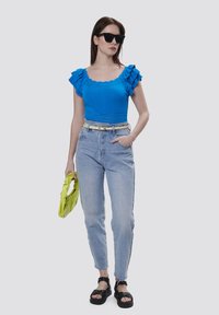 Blauses Stricktop mit Rüschensleeves, hochgeschnittene helle Jeans und schwarze Sandalen. Als Accessoires dienen eine limegrüne Clutch und schwarze Sonnenbrille.