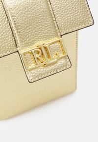 Angolo di una borsa in pelle dorata con texture, dotata di una chiusura a pattina con il logo dorato "RL" su uno sfondo bianco.