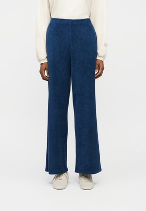 Osoba oblečená v voľných tmavomodrých nohaviciach s elastickým pásom a bielych teniskách, stojaca na jednoduchom pozadí.