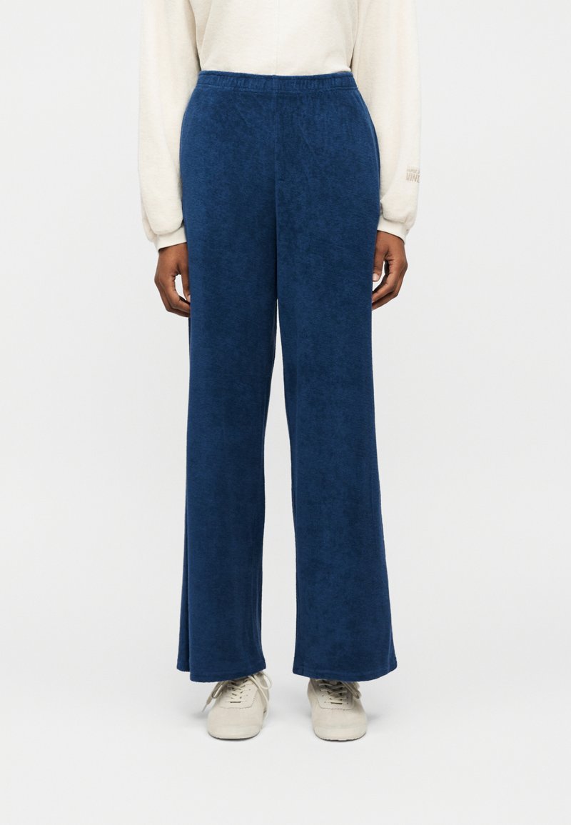 Άτομο που φοράει φαρδιά, σκούρα μπλε παντελόνια με ελαστική ζώνη στη μέση και λευκά αθλητικά παπούτσια, στέκεται μπροστά σε απλό φόντο.