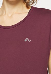 Burgundy idrottstopp med rund halsringning, korta ärmar och en grå cirkulär logotypaccents på vänster bröst. Slät tygtextur.