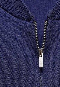Tejido de punto azul marino con cuello de canalé y cremallera metálica. La cremallera tiene un tirador plateado y dientes expuestos.