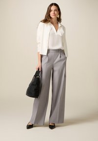 Mujer con blusa blanca y cárdigan, pantalones anchos grises y tacones negros, sosteniendo un bolso negro sobre un fondo beige.