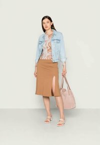 Casaco de ganga, blusa ajustada com padrão, saia castanha de comprimento médio com racha lateral, sandálias brancas e uma mala de Tote rosa claro. Fundo neutro.