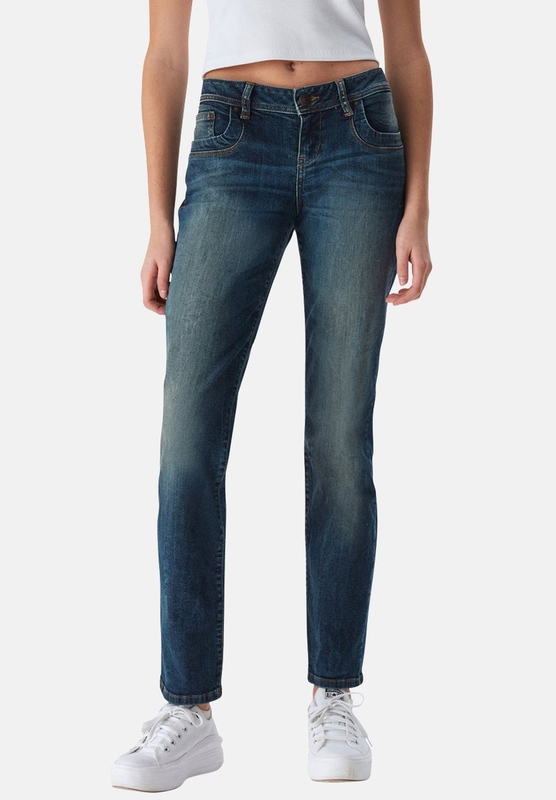 Jeans de mezclilla de color azul oscuro desgastado, diseño de pierna recta, con bolsillos frontales, cierre de botón y textura desgastada. Combinados con zapatillas blancas.