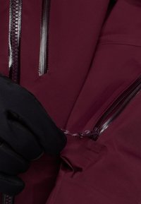 Vinröd jacka med glansiga svarta blixtlås och dragsko. Svarta handskar greppar tyget och visar upp dess struktur och designdetaljer.