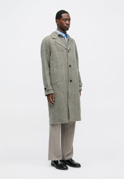 Sivi herringbone kaput s izrezanim ovratnikom, velikim gumbima i bočnim džepovima, nosi se preko bež košulje i sivih hlača. Crne cipele.