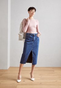 Helles rosa strukturiertes Langarmoberteil, hochtaillierter blauer Jeansrock mit Schlitz vorne und weiße Absatzschuhe. Modell posiert mit einer Jacke über der Schulter.