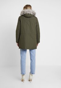 Olivově zelená parka s kapucí lemovanou umělou kožešinou, má mírně stažený pas, dlouhé rukávy a rovný střih. K ní jsou nošeny světle modré džíny.