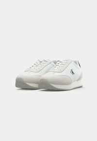 Witte en lichtgrijze lage sneakers met veters, gestructureerde rubberen zolen, en zwart "CK" logo aan de zijkant, getoond vanuit een voorlinkse hoek.