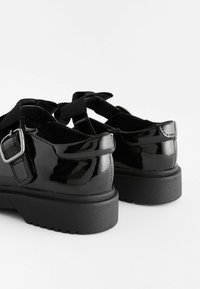 Chaussures Mary Jane en cuir verni noir avec semelles épaisses en caoutchouc et grandes brides à boucle, présentées de dos sur fond blanc.