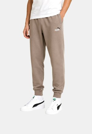 Persona con pantalones deportivos beige de Puma y zapatillas blancas Puma con raya negra, de pie con una mano en el bolsillo sobre un fondo liso.