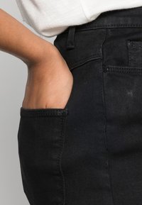Pantalon en denim noir avec une poche visible ; gros plan sur une main reposant dans la poche. Le tissu semble lisse avec une texture minimale.
