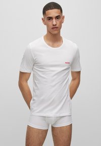T-shirt en coton blanc avec un col rond et des manches courtes, présentant un petit logo rouge "HUGO" sur la poitrine gauche. Le modèle porte des sous-vêtements blancs.