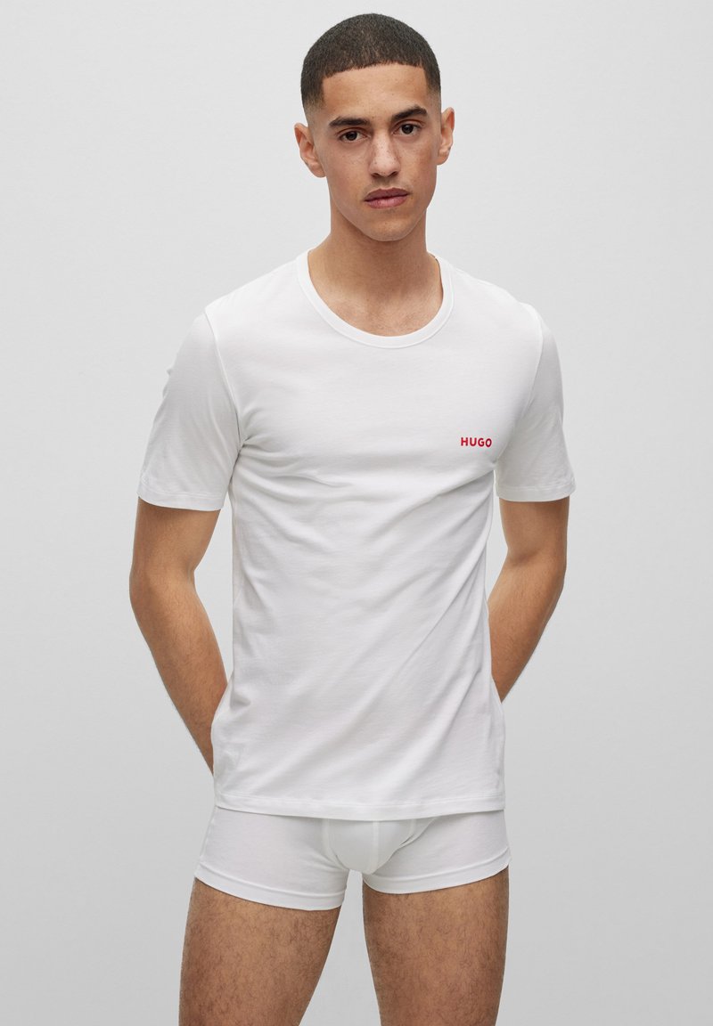 T-shirt en coton blanc avec un col rond et des manches courtes, présentant un petit logo rouge "HUGO" sur la poitrine gauche. Le modèle porte des sous-vêtements blancs.