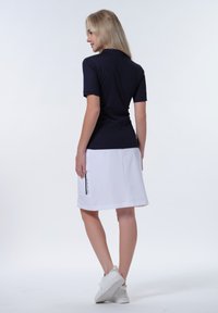 Marineblaues Poloshirt mit kurzen Ärmeln, kombiniert mit einem weißen Rock mit einer Seitenasche und schwarzen Akzenten, getragen mit weißen Turnschuhen.