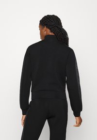 Zwarte sportjack met hoge kraag en ribgebreide manchetten. Beschikt over een gladde stof en een logo-accent dat langs de mouwen loopt.