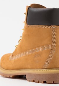 Botte de travail en cuir marron clair avec col rembourré noir, lacets jaunes et rouges, coutures blanches visibles et semelle en caoutchouc marron robuste.