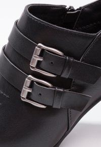 Botín negro de piel suave con dos detalles de hebillas plateadas, que presenta una forma redondeada y costuras visibles.