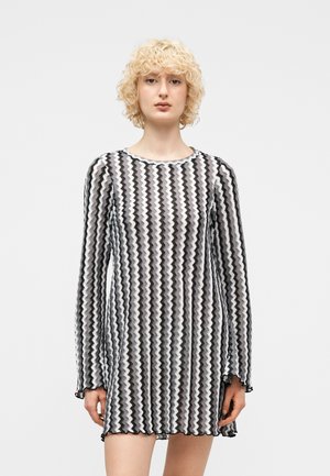 Ung person med krøllet blondt hår iført en sort og hvid zigzag-mønsteret langærmet kjole mod en ensartet lys baggrund.