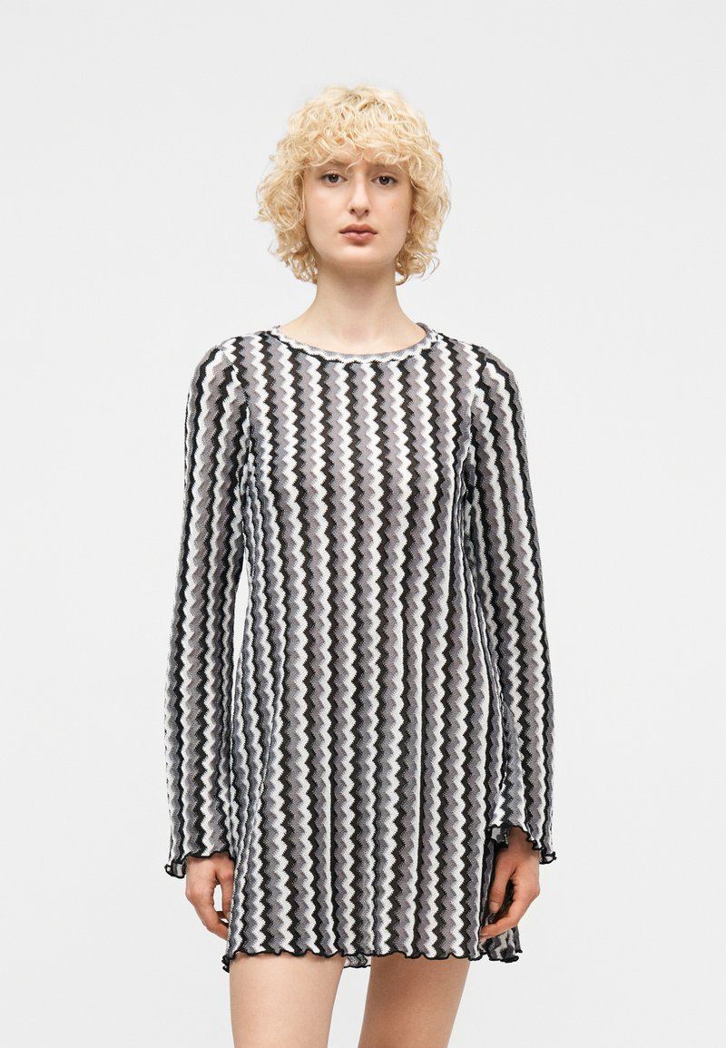 Νεαρό άτομο με σγουρά ξανθά μαλλιά, που φορεί μακρυμάνικο φόρεμα με μαύρο και λευκό ζιγκ-ζαγκ σχέδιο, μπροστά από ένα απλό ανοιχτό φόντο.