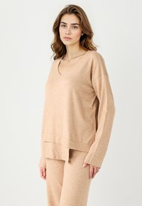 Top beige a scollatura a V realizzato in tessuto morbido, con maniche lunghe, una vestibilità rilassata e un sottile motivo testurizzato. Include un design a orlo multilivello.