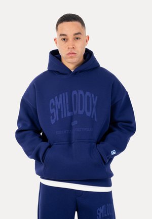 Ung mand iført en mørkeblå Smilodox-hættetrøje og matchende joggingbukser med hænder i forlommerne foran en ensfarvet baggrund.