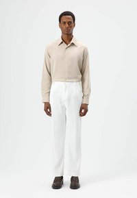 Lichtbeige shirt met lange mouwen en een knoopsluiting, gecombineerd met witte pantalon met fijne streepjes en stevige donkerbruine schoenen.