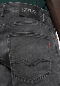 Κοντινό πλάνο της πίσω τσέπης μαύρων τζιν με μαύρη ραφή και ετικέτα Replay Blue Jeans στο ζωνάκι.