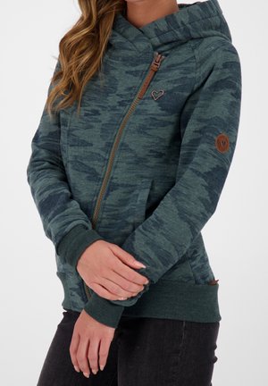 Femme portant un sweat à capuche vert à motif camouflage avec une fermeture éclair asymétrique, un logo cœur sur la poitrine et un patch rond sur la manche.
