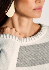 Nahaufnahme des unteren Gesichts einer Frau, ihres Halses und ihrer Hand, die einen cremefarbigen und grauen Strickpullover sowie große cremefarbene Ohrringe trägt.