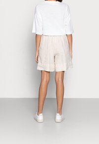 Pantaloni corti in lino beige con vestibilità rilassata e orlo arrotolato, abbinati a una T-shirt bianca a maniche corte e sneakers bianche con logo.