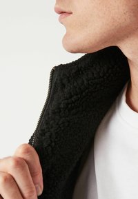 Černá fleecová bunda s límcem, který má strukturovaný, nadýchaný povrch a zapínání na zip. Pod ní je bílé tričko, částečně viditelná ruka na límci.
