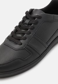 Baskets noires en cuir lisse avec des trous de ventilation, bout rond, lacets plats noirs et une discrète marque Calvin Klein sur le côté.