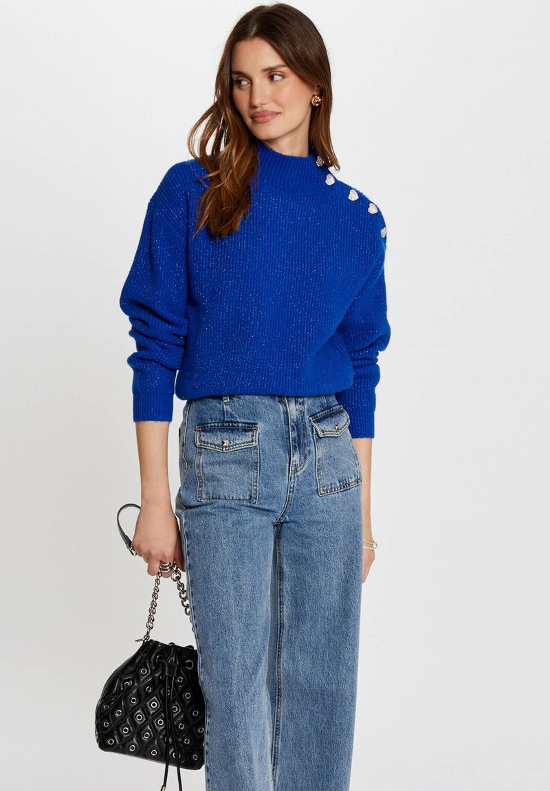 Pull en tricot bleu avec une texture côtelée et un col haut, orné de boutons argentés sur une épaule ; associé à un jean en denim taille haute.