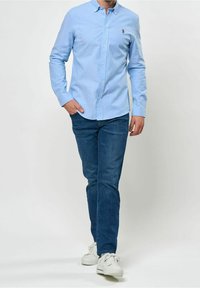 Man med ljusblå skjorta, mörkblå jeans och vita sneakers som går med en hand i fickan mot vit bakgrund.