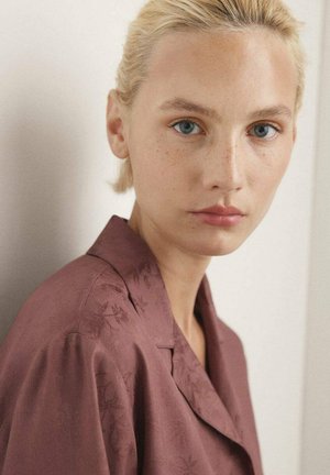 Chemise marron avec une texture lisse et un motif floral, dotée d'un col à encoches et de manches tombantes, portée par une personne à la peau claire.