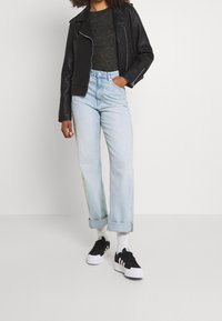Schwarze Lederbikerjacke, graues, figurbetontes Hemd, hellblaue Jeans mit aufgerollten Säumen, schwarz-weiße Sneakers mit drei Streifen, einfarbiger Hintergrund.