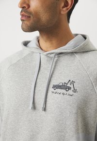 Grå hættebluse med lysegrå detaljer og en broderet design af en lastbil med en løftarm samt teksten "Vi løfter det hele. Store & små."