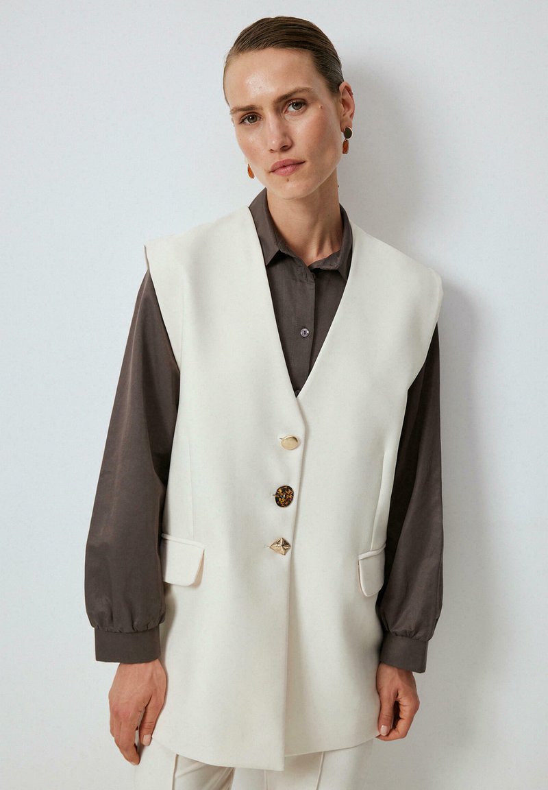 Chaleco blanco sin mangas con cuello de muesca, cuatro botones (dos dorados, uno de carey y uno plateado), y bolsillos laterales, usado sobre una blusa gris oscura.