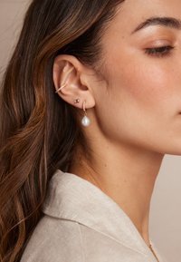 Pendientes de oro con una perla caída, acompañados de un pequeño charm cuadrado. Los grilletes de las orejas están adornados con diminutas piedras preciosas.