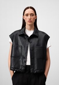 Gilet in pelle nera con collo, design senza maniche, due tasche frontali e chiusura a bottoni, indossato sopra una t-shirt bianca.