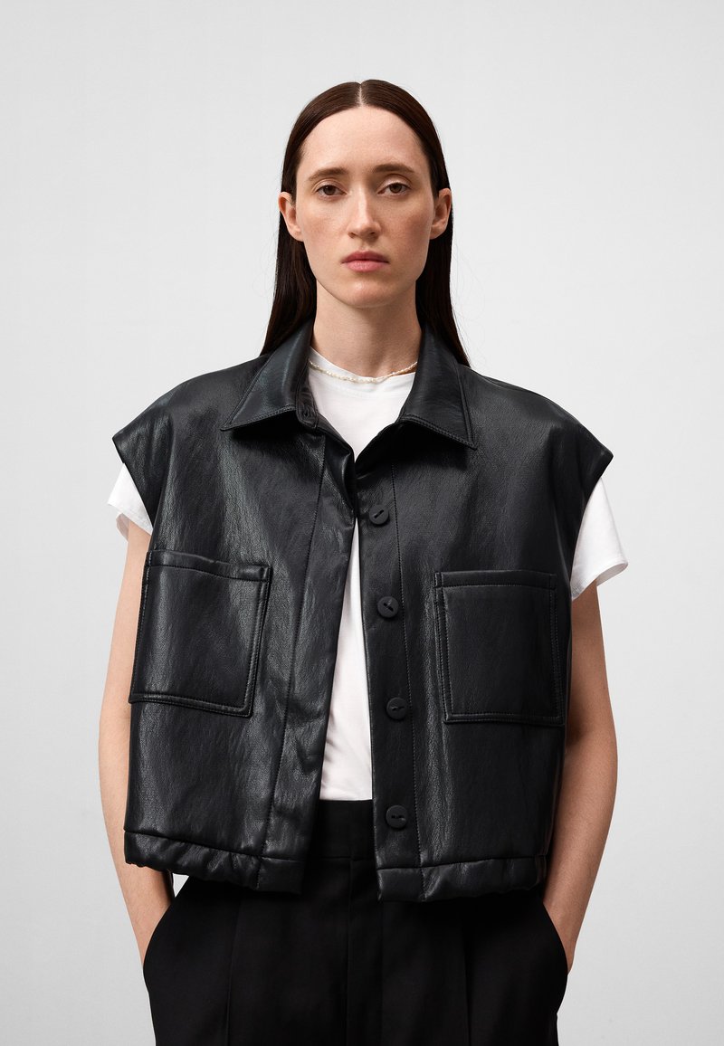 Gilet in pelle nera con collo, design senza maniche, due tasche frontali e chiusura a bottoni, indossato sopra una t-shirt bianca.