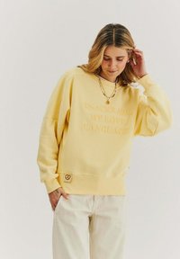 Žltý nadrozmerný sveter s vyšívaným textom "SNACKS ARE MY LOVE LANGUAGE," manžety s rebrovaním a látkovou nášivkou s grafikou prelomeného chleba.