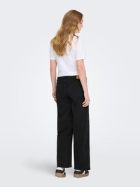 Jeans neri a gamba larga con tasche posteriori, abbinati a una t-shirt bianca a maniche corte. I capelli cadono in onde morbide, mostrando uno stile semplice e casual.