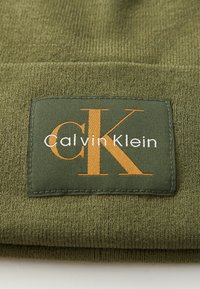Beanie verde oliva realizzato in tessuto texturizzato, caratterizzato da una toppa rettangolare con la scritta "Calvin Klein" e "CK" in lettere arancioni e bianche.